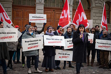 La présidente d'Unia Vania Alleva tient un discours devant un groupe de militant-e-s aux bouches scotchées tenant des panneaux protestant contre les licenciements abusifs.
