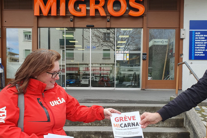 Gewerkschafterin in roter Jacke mit Unia-Logo verteilt einem Passanten ein Flugblatt zum Micarna-Streik vor einer Migros-Filiale.