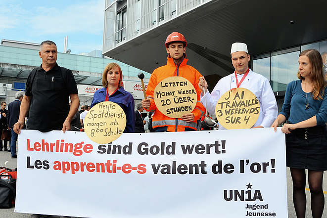 5 syndicalist-e-s avec affiche et plakettes: J'ai travaillé 3 weekends sur 4.Lehrlinge sind Gold wert. Heute Spätdienst, morgen ab 8.00 Schule. Letzten Montat 20 Überstunden. 