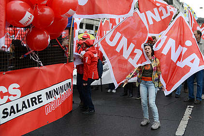 Une femme dans une manifestation est entourée de drapeaux du syndicat Unia. à sa gauche, une pancarte demandant un salaire minimum.