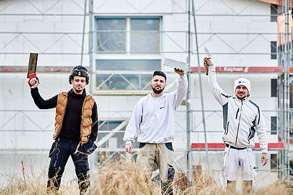 Drei Arbeiter im Aussengewerbe erheben ihre Werkzeuge vor einem Gerüstbau