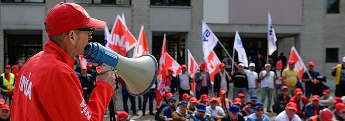 Un secrétaire syndical d'Unia parle devant des travailleurs du second œuvre romand assis et debout, avec des drapeaux