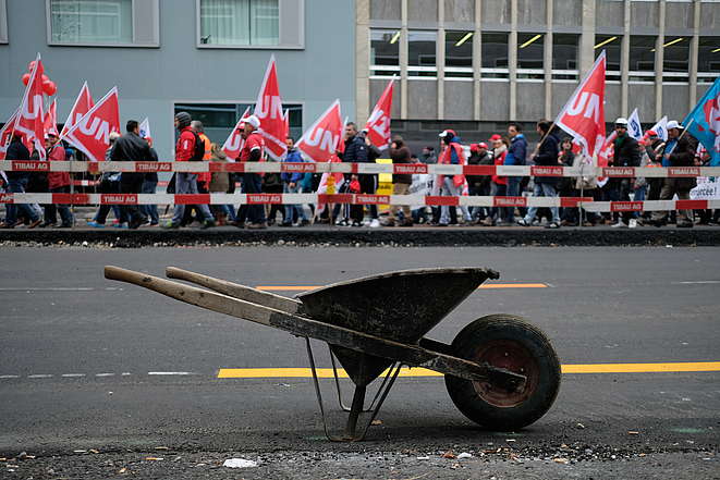 Una carriola vuota lascia intravedere un corteo di lavoratori edili.