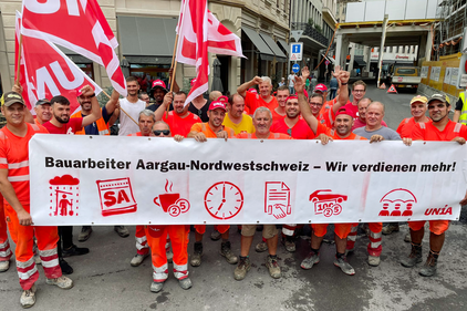 Bauarbeiter in organger Arbeitskleidung mit Transpi: Bauarbeiter Aargau-Nordwestscheiz - Wir verdienen mehr!
