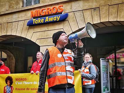 Ein Unia-Mann mit einem Megafon und Menschen mit einem Transparent vor einem Migros Take Away